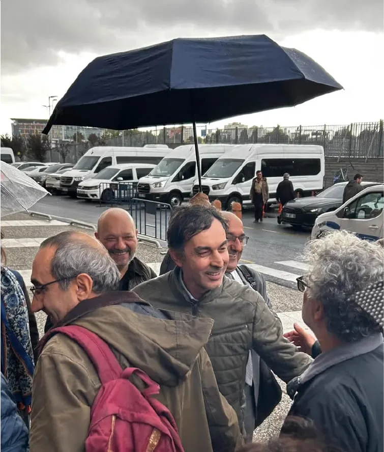Şemsiyeli, sırt çantalı bir grup insan binanın önünde bir araya gelmiş, arkada beyaz minibüsler görünüyor.