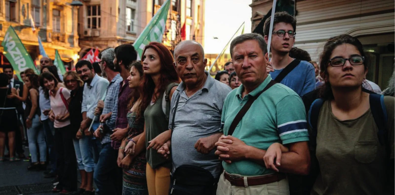 Sokakta kol kola girmiş, bayraklar taşıyan bir kalabalık.