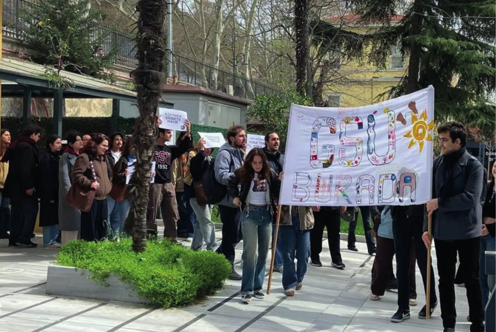 Öğrenciler kampüs bahçesinde toplanmış, biri “GSÜ BURADA” yazılı el yapımı pankartı taşıyor.