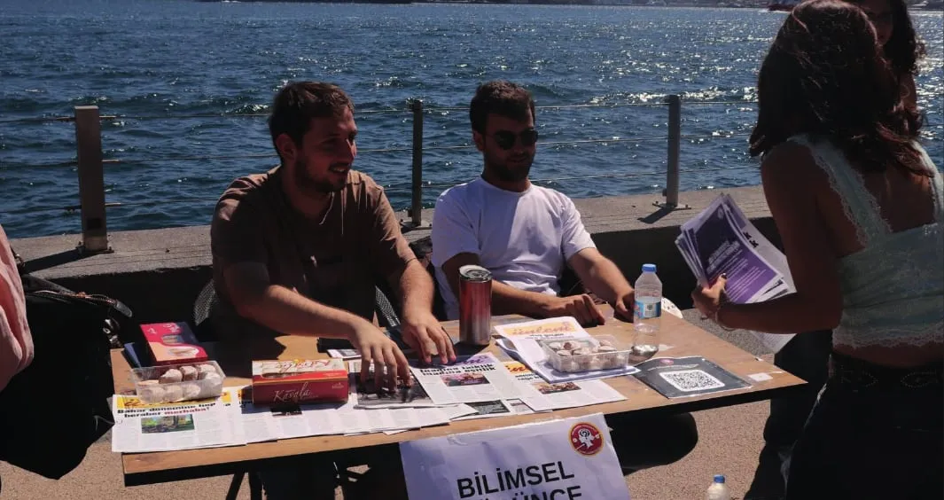 Deniz kıyısında Bilimsel Düşünce Kulübü masasında oturan iki öğrenci ve broşür tutan bir ziyaretçi.