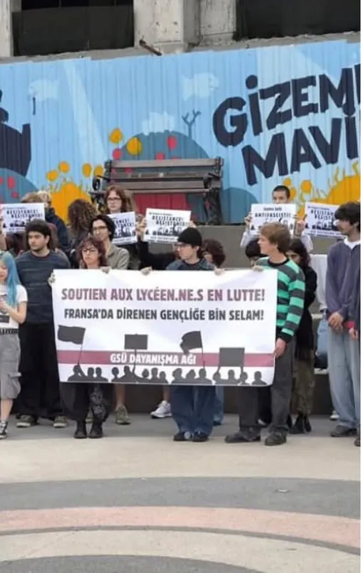 Öğrenciler “Fransa’da direnen gençliğe bin selam!” yazılı GSÜ Dayanışma Ağı pankartıyla bir meydanda duruyor.